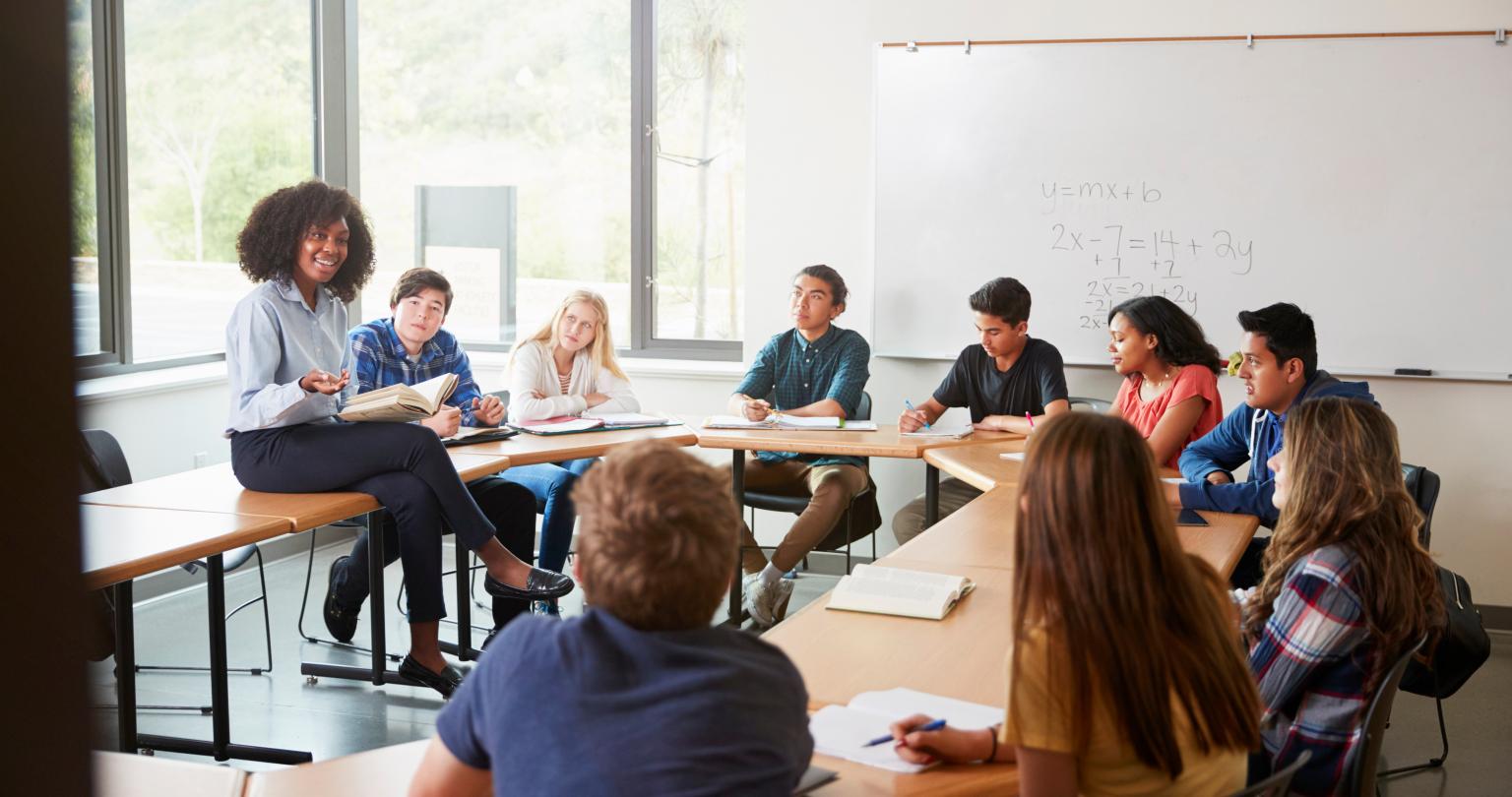 Female High School Tutor Sitting At Table With Pupils Teaching Maths Class