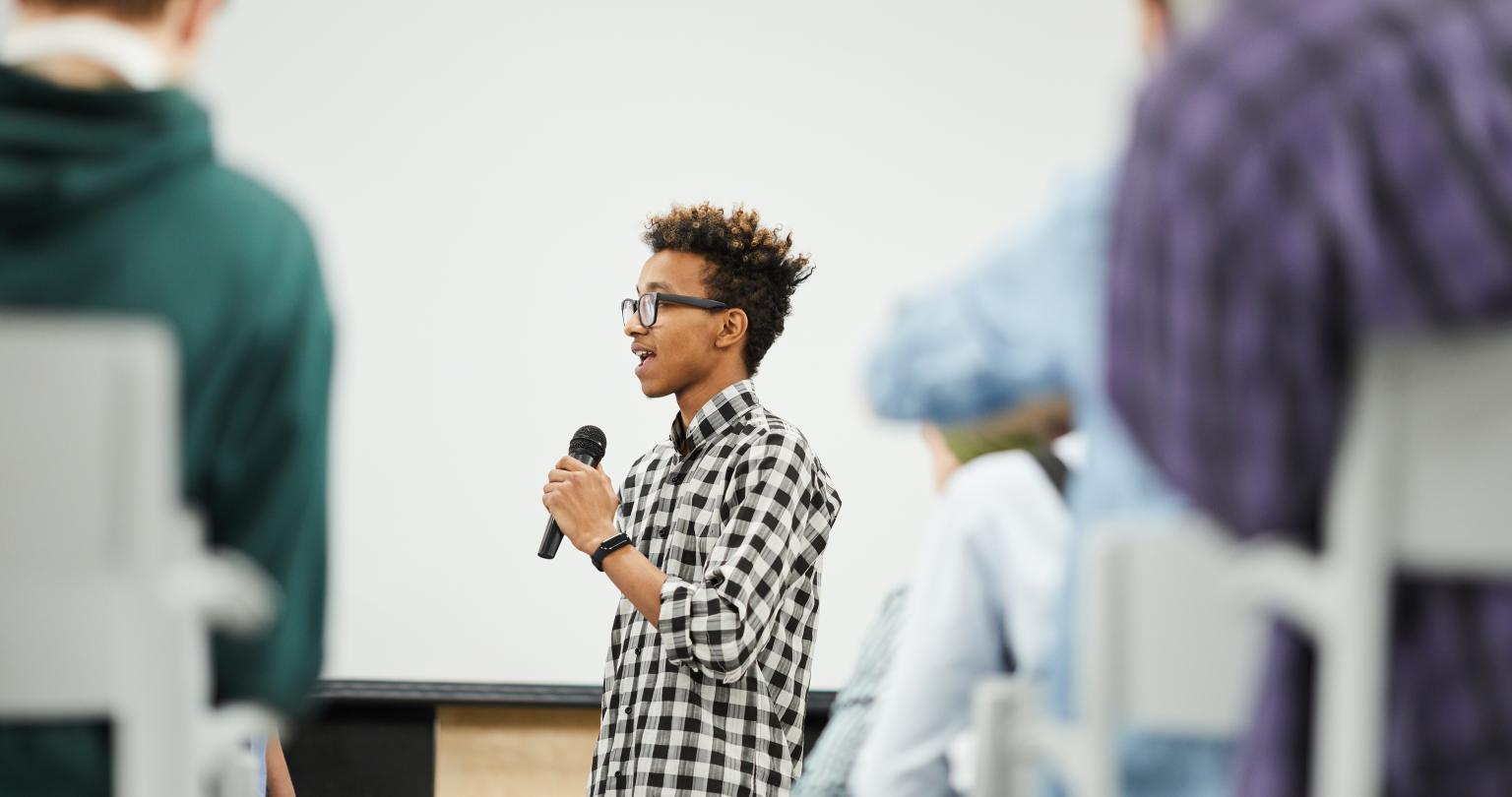 Cheerful confident young black student with Afro hairstyle standing in convention center room and presenting his startup project at conference