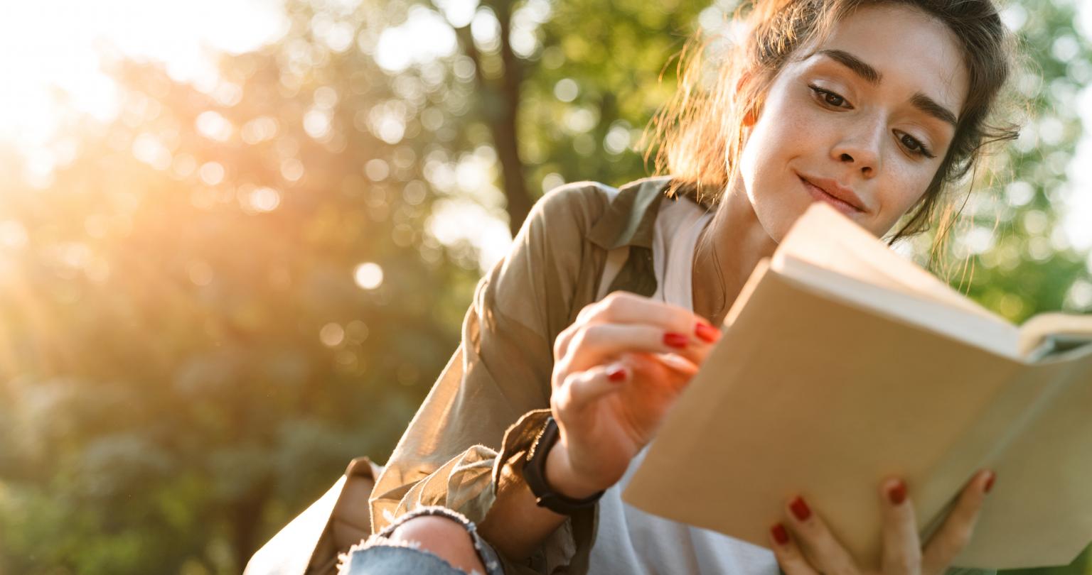 Image of young woman wearing casual clothes smiling and reading book while walking in green park