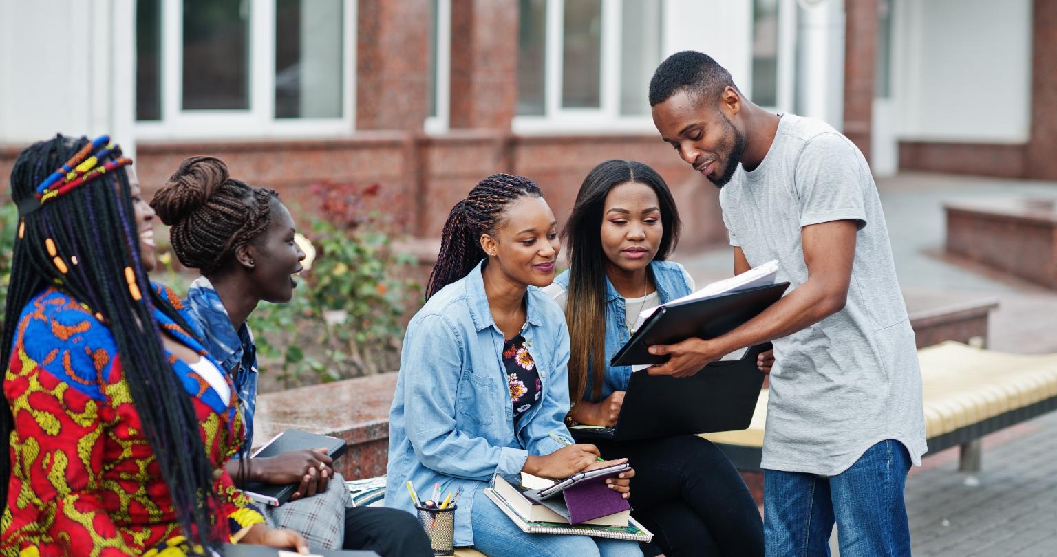 Group of five african college students spending time together on campus at university yard. Black afro friends studying at bench with school items, laptops notebooks.