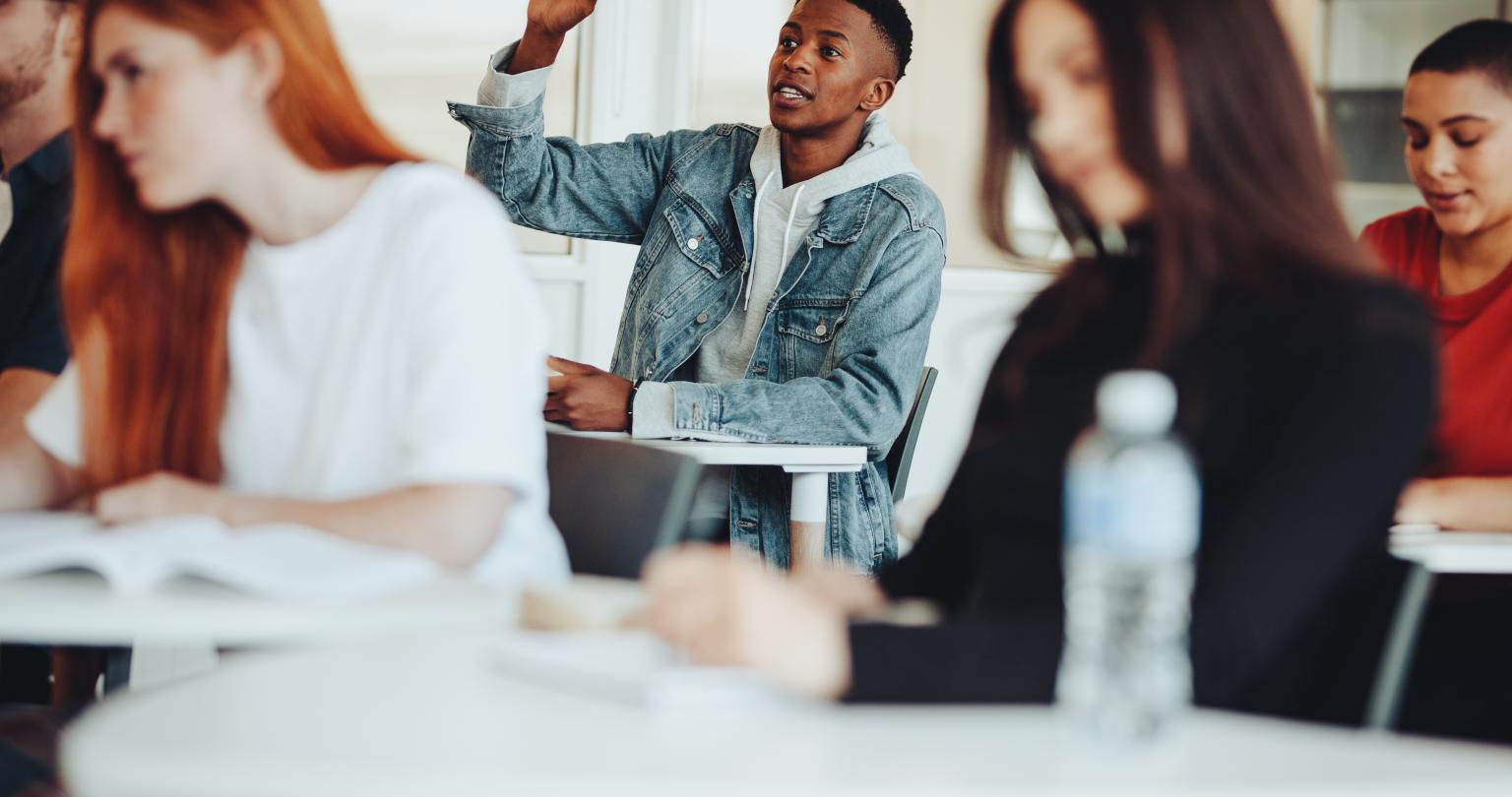 Male student sitting in the class and raising hand to ask a question during lecture. College student asking a query to the lecturer in classroom.