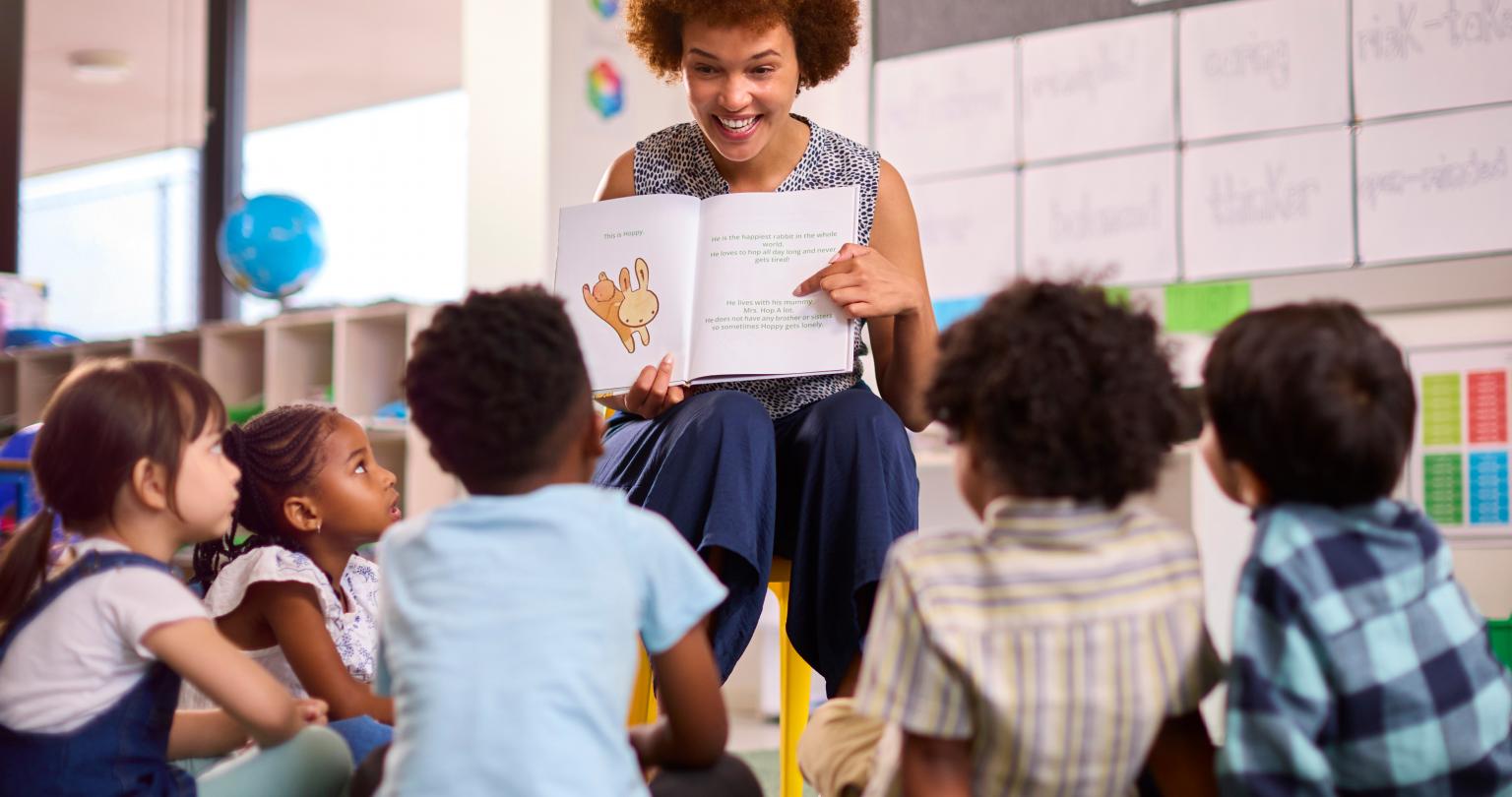 Female Teacher Reads To Multi-Cultural Elementary School Pupils Sitting On Floor In Class At School