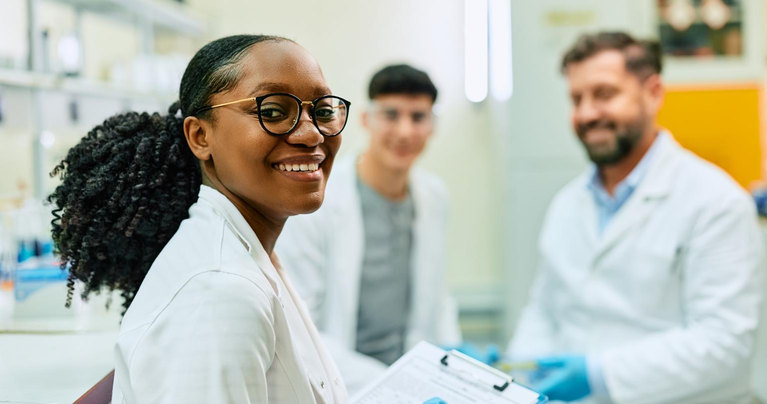 Happy black biochemist working with her colleagues in lab and looking at camera.