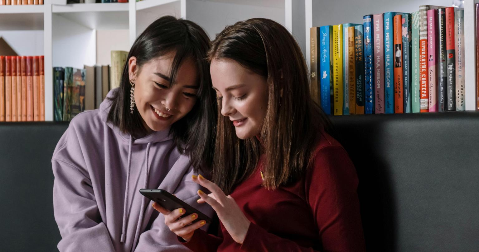 Two teenaged girls sitting with their backs to a bookcase and looking down at one phone.