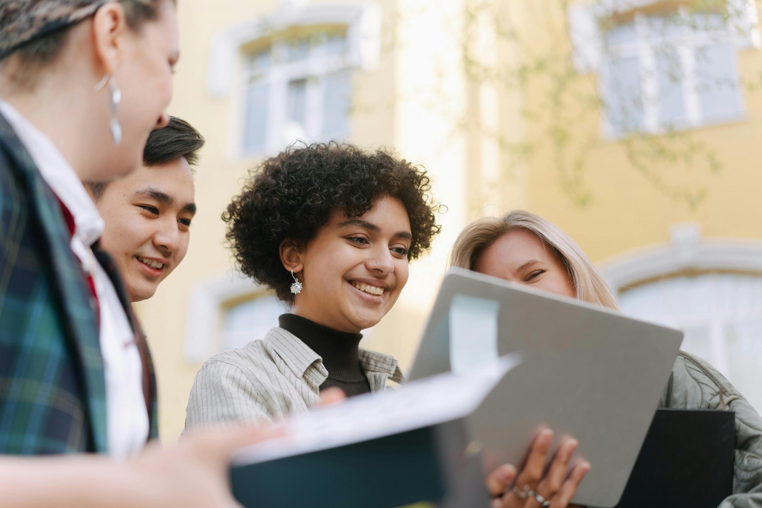 Diverse group of 4 teenage students smiling and looking at a single computer screen outside.