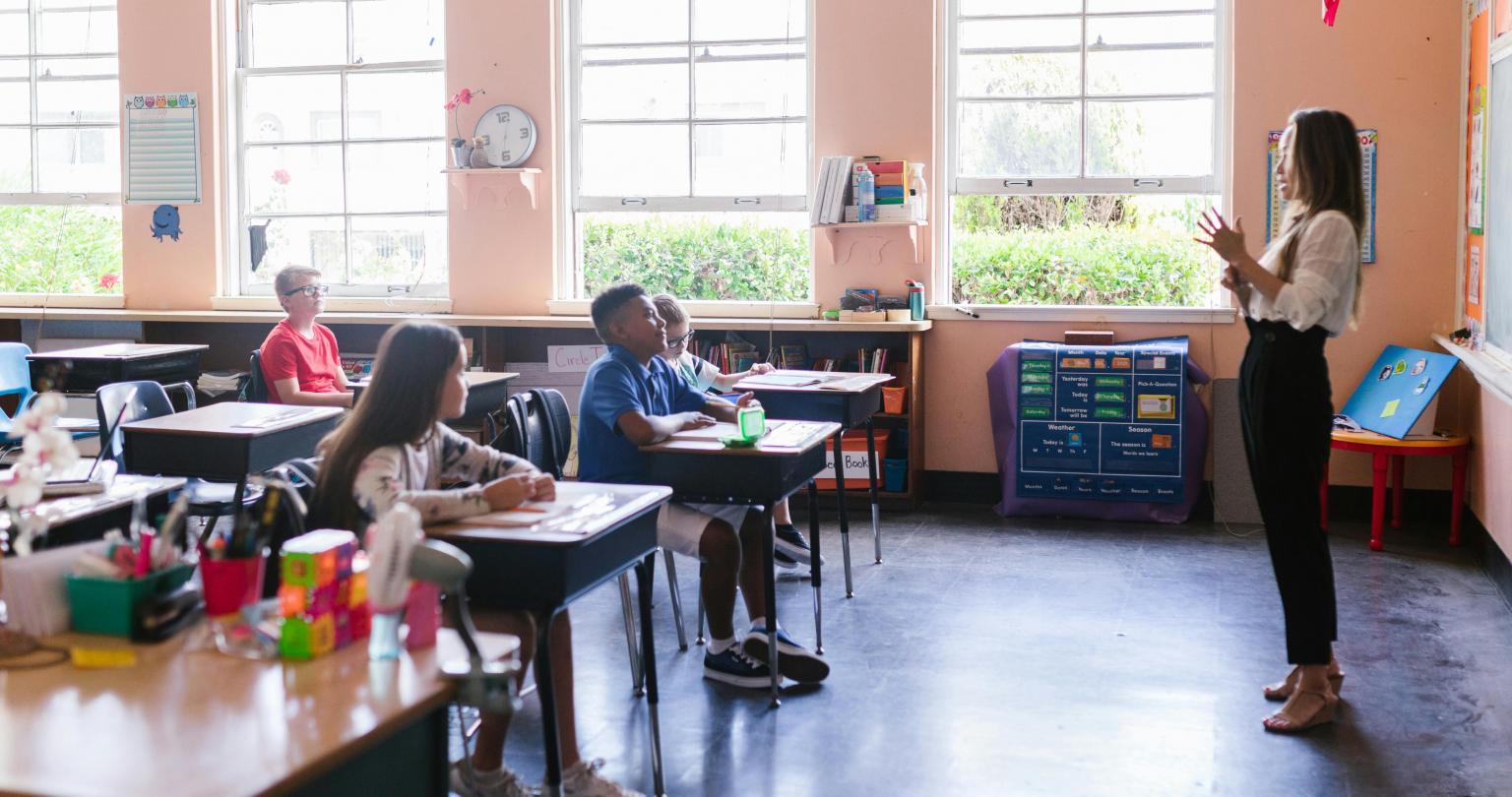 Teacher speaking at the front of the room with elementary kids sitting at their tables.